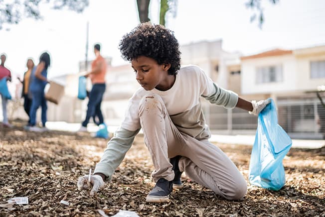 Young boy wearing rubber gloves and holding a trash bag while picking up a soda can in a residential neighborhood